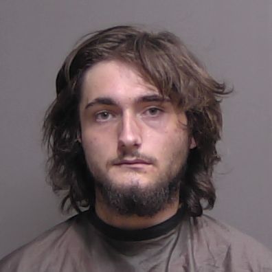 A young man with long brown hair and a partial beard looks at the camera with a neutral expression, wearing a dark cape against a plain gray background A young man with long brown hair and a partial beard looks at the camera with a neutral expression, wearing a dark cape against a plain gray background