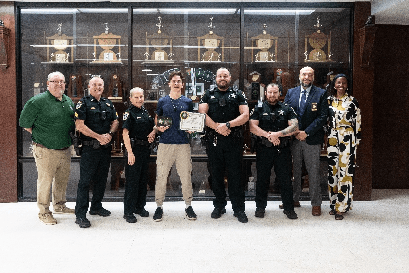 A group of eight people, including law enforcement officers and civilians, stand in front of a trophy case One young man in the center holds a certificate and poses with others smiling beside him