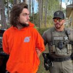 A man in an orange hoodie with an anime logo is being escorted outdoors by a sheriff’s deputy in tactical gear and sunglasses Trees and a building are visible in the background