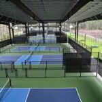 A covered outdoor facility with multiple blue and green pickleball courts separated by black fencing, surrounded by trees and open land