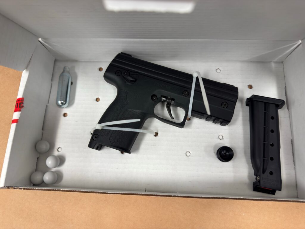 A black airsoft pistol, extra magazine, CO cartridge, round pellets, and a small black cap, all secured in a white cardboard box with zip ties