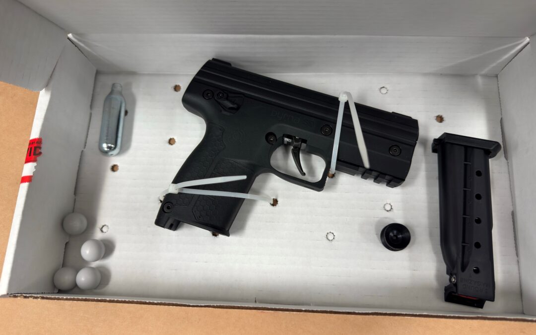 A black airsoft pistol, extra magazine, CO cartridge, round pellets, and a small black cap, all secured in a white cardboard box with zip ties