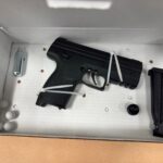 A black airsoft pistol, extra magazine, CO cartridge, round pellets, and a small black cap, all secured in a white cardboard box with zip ties