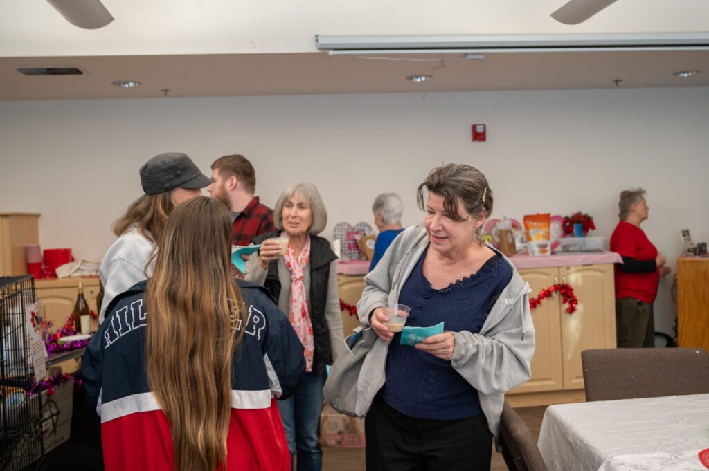 A group of people mingle and chat at an indoor gathering Some hold drinks, and there are decorations and items on tables and shelves in the background One woman in a gray hoodie talks and smiles in the foreground