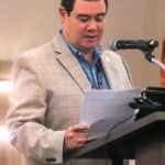 A man in a light plaid blazer stands at a podium, reading from papers, with a microphone in front of him He appears to be speaking or presenting at an indoor event or conference