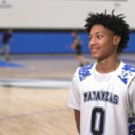 A young basketball player in a white "Matanzas" jersey with the number stands on an indoor court, smiling In the background, other players practice on the basketball court
