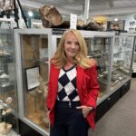 A woman with long blonde hair in a red blazer stands in a museum exhibit filled with fossils and rocks displayed in glass cases She is smiling and has one hand in her pocket