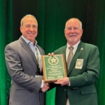 Two smiling men in suits stand in front of a green backdrop, shaking hands while one holds a plaque with a star emblem Both wear badges, and the man in the green blazer is presenting the award