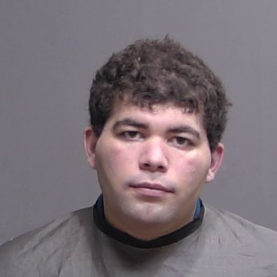 A man with short, curly dark hair is facing the camera against a plain, neutral background, wearing a dark colored protective cover over his shoulders