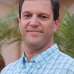 A man with short brown hair wearing a blue and white plaid shirt stands outdoors in front of palm trees and a peach colored building