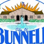 Logo for the City of Bunnell featuring a building with an American flag, a fountain in front, orange sun rays, green trees, and "City of Bunnell" written in bold blue letters at the bottom