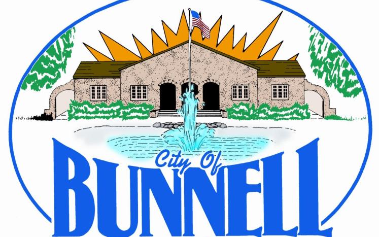 Logo for the City of Bunnell featuring a building with an American flag, a fountain in front, orange sun rays, green trees, and "City of Bunnell" written in bold blue letters at the bottom