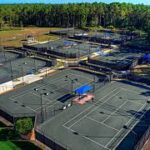Aerial view of multiple outdoor tennis courts surrounded by trees, with blue shade structures and fencing around each court The courts are arranged in rows and the area appears to be part of a large sports complex