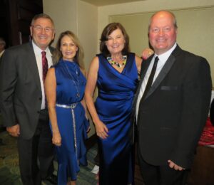 Four adults stand together at an indoor event, dressed in formal attire The two women in the middle wear blue dresses, while the men on either side wear dark suits and ties All are smiling at the camera Four adults stand together at an indoor event, dressed in formal attire The two women in the middle wear blue dresses, while the men on either side wear dark suits and ties All are smiling at the camera
