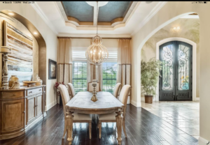 Elegant dining room with a wooden table, six chairs, a sideboard, large windows with drapes, a modern chandelier, dark wood flooring, and an arched entryway leading to a grand double front door Elegant dining room with a wooden table, six chairs, a sideboard, large windows with drapes, a modern chandelier, dark wood flooring, and an arched entryway leading to a grand double front door