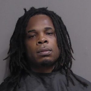 A man with medium length dreadlocks and a serious expression is wearing a dark cape, standing against a plain gray background