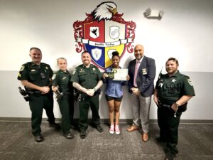 A group of six people, including four uniformed officers and two civilians, stand smiling together One woman in the center holds a certificate They pose in front of a wall with a large colorful crest and the words "Buddy Taylor A group of six people, including four uniformed officers and two civilians, stand smiling together One woman in the center holds a certificate They pose in front of a wall with a large colorful crest and the words "Buddy Taylor