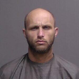 A man with a shaved head and short beard wearing a gray shirt stares at the camera with a serious expression against a plain gray background