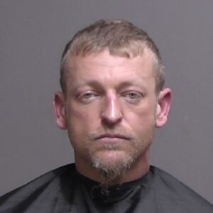 A man with short light brown hair, light facial hair, and a neutral expression is shown against a plain background, wearing a dark drape often used for mugshots or identification photos A man with short light brown hair, light facial hair, and a neutral expression is shown against a plain background, wearing a dark drape often used for mugshots or identification photos