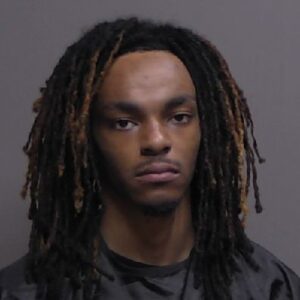 A young Black man with long dreadlocks, wearing a dark shirt, photographed against a gray background in what appears to be a mugshot style photo