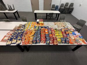 A large assortment of packaged snacks, including chips, ramen noodles, and cookies, neatly arranged on two tables in a room with gray chairs, white walls, and stacked furniture in the background