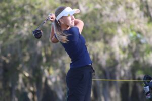 A woman in a navy shirt and white visor swings a golf club outdoors, with blurred green trees in the background A woman in a navy shirt and white visor swings a golf club outdoors, with blurred green trees in the background