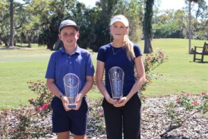 Two young golfers, a boy and a girl, stand outside on a golf course, each holding a large clear trophy and smiling at the camera Trees and grass are in the background Two young golfers, a boy and a girl, stand outside on a golf course, each holding a large clear trophy and smiling at the camera Trees and grass are in the background