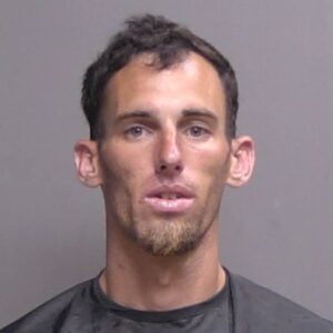 A mugshot style photo of a lean white man with short dark hair, light stubble, and a neutral expression, wearing a dark shirt against a gray background