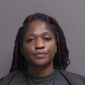 A woman with braided hair wearing a dark top poses against a plain, neutral colored background, looking directly at the camera with a neutral expression
