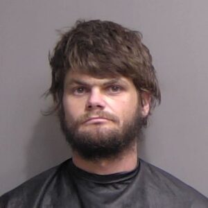 A man with medium length brown hair and a beard, wearing a dark cape or smock, stands against a plain, light colored background, looking directly at the camera with a neutral expression