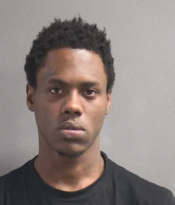 A young man with short, curly hair and a serious expression faces the camera against a plain, gray background He is wearing a black T shirt
