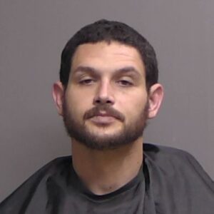 A man with short dark hair and a beard is wearing a black drape, posing against a plain gray background He is facing the camera with a neutral expression A man with short dark hair and a beard is wearing a black drape, posing against a plain gray background He is facing the camera with a neutral expression