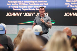 A man with tattoos, wearing a gray polo and khaki pants, speaks into a microphone on stage before an audience A large display behind him shows his name, Johnny "Joey" Jones, and mentions his professional credentials