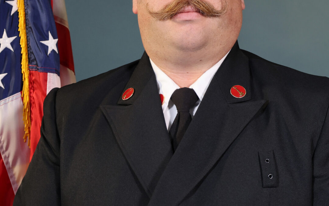 A man in a formal dark uniform with brass buttons stands in front of an American flag He has short dark hair, glasses, and a styled mustache, and is posing against a plain blue background
