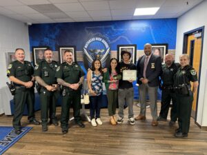 A group of police officers, school staff, and three civilians stand together at Matanzas High School One young man holds a certificate, smiling, with others gathered and smiling for the photo in a hallway A group of police officers, school staff, and three civilians stand together at Matanzas High School One young man holds a certificate, smiling, with others gathered and smiling for the photo in a hallway