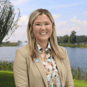 A woman with long blonde hair wearing a tan blazer and a floral blouse smiles while standing outdoors near a lake, with trees, grass, and a partly cloudy sky in the background
