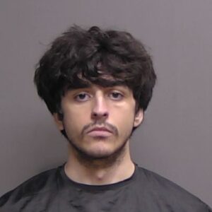 A young man with curly dark hair and a mustache is wearing a black garment, standing against a plain, neutral background in a Flagler County mugshot style photo