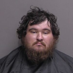 A man with a thick brown beard and curly hair is facing the camera with a neutral expression, wearing a dark garment against a plain background