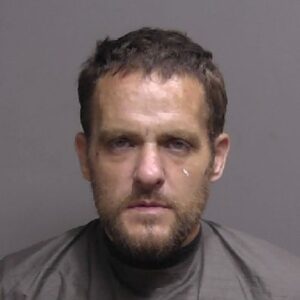 A man with short brown hair and a beard is wearing a gray garment and looking straight at the camera against a plain gray background