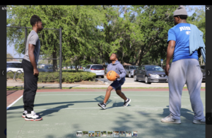 A young boy prepares to shoot a basketball on an outdoor court while two adults watch, one standing on each side Cars and trees are visible in the background under a clear blue sky