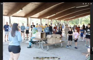A group of people, including children and adults, stand under a pavilion listening to a woman speak A costumed mascot sits at a picnic table with cardboard boxes on it Trees are visible outside the pavilion A group of people, including children and adults, stand under a pavilion listening to a woman speak A costumed mascot sits at a picnic table with cardboard boxes on it Trees are visible outside the pavilion