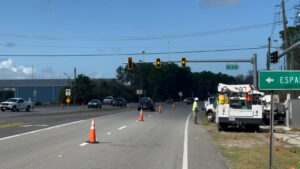 Traffic cones and a utility truck are on the side of a multi lane road near a traffic light Workers in safety vests are present A green road sign points right to "ESPA " Several cars are driving ahead under a blue sky