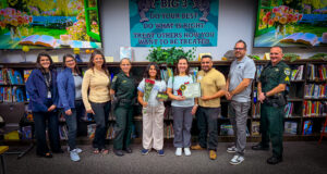 A group of ten people, including police officers and civilians, stand smiling in a library One person in the center holds flowers and a certificate Behind them is a colorful sign with inspirational quotes A group of ten people, including police officers and civilians, stand smiling in a library One person in the center holds flowers and a certificate Behind them is a colorful sign with inspirational quotes