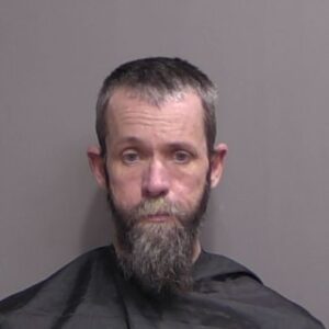 A middle aged man with short dark hair and a long gray beard faces the camera against a plain gray background, wearing a black garment similar to a barber’s cape