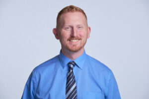 A man with short red hair and a trimmed beard smiles at the camera He is wearing a bright blue dress shirt and a black and gray striped tie, standing against a plain light background