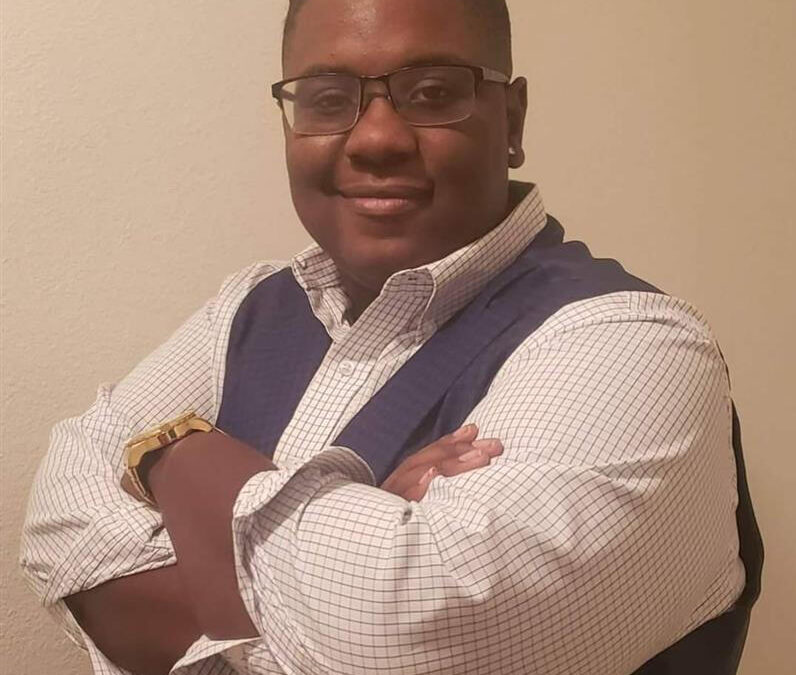 A person wearing glasses, a checked button up shirt, and a blue vest stands with arms crossed, smiling at the camera against a plain beige background