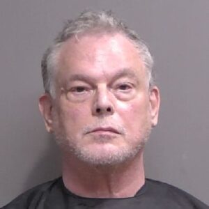 A middle aged man with short gray hair and a trimmed beard is facing the camera against a plain background, wearing a dark garment typically used for mugshots