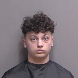 A young person with short, curly dark hair and light skin looks directly at the camera against a plain gray background, wearing a dark cape typically used for mugshots or official photos