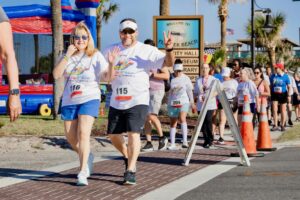 Two people wearing numbered race bibs smile and wave as they walk in a fun run event with other participants behind them Palm trees, orange cones, and a welcome sign are visible in the background Two people wearing numbered race bibs smile and wave as they walk in a fun run event with other participants behind them Palm trees, orange cones, and a welcome sign are visible in the background
