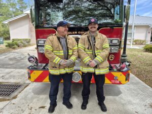 Two firefighters in tan jackets and navy pants stand smiling in front of a red fire truck, outdoors on a sunny day with trees and buildings in the background